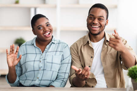 Happy African Couple Waving Hand Smiling To Camera Sitting Indoors