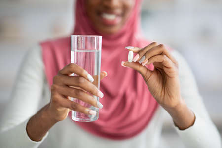 Black Woman In Hijab Taking Supplement Pill And Holding Glass Of Waterの写真素材