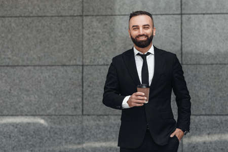 Happy entrepreneur in black suit drinking coffee to go during break, standing near office center wall, copy spaceの写真素材