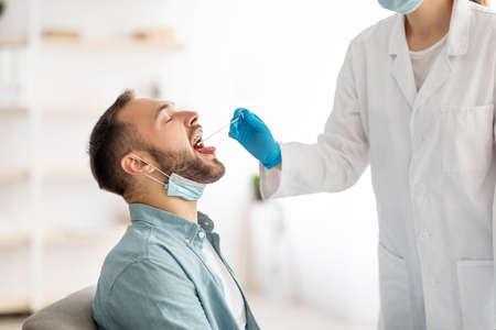 Doctor making oral PCR test for young man at clinic. Millennial guy undergoing diagnostic procedure
