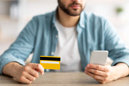 Shopping at home. Closeup of young guy holding credit card and smartphone, making online purchase at...