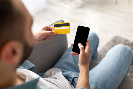 Ordering goods online. Unrecognizable young guy with credit card and smartphone shopping on web at...