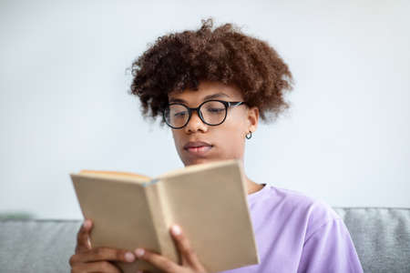 Focused African American teenager reading book, enjoying captivating story at homeの写真素材