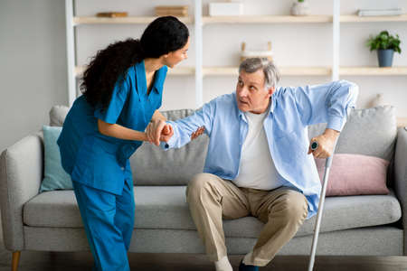 Millennial caregiver helping senior man with crutches to get up from couch at retirement home