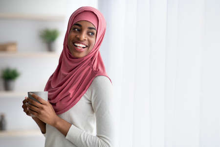 Happy Black Muslim Woman In Hijab Drinking Coffee Near Window At Home