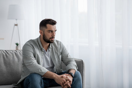 Thoughtful serious bearded millennial male sit on sofa alone at homeの写真素材