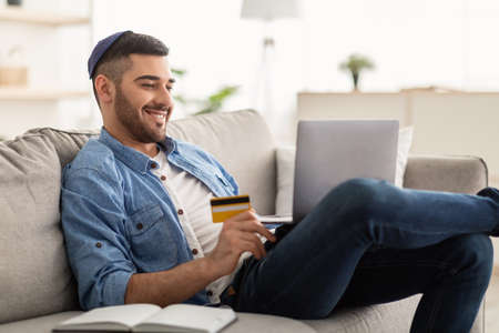 Smiling jewish guy using laptop and credit card at home