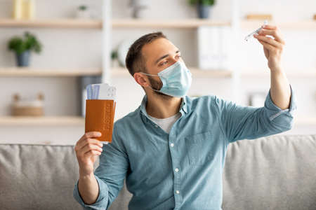 Coronavirus travel ban. Upset young guy in face mask holding passport and plane tickets, playing with toy plane at homeの写真素材