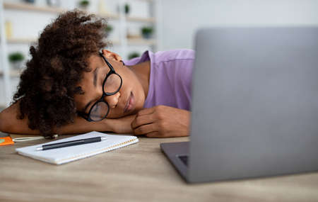 Tired black teenager sleeping on table in front of laptop computer at home, panoramaの写真素材