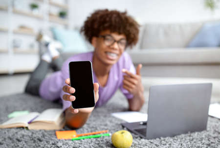Black teenager lying on floor with laptop, pointing at smartphone with blank screen at home, mockup...