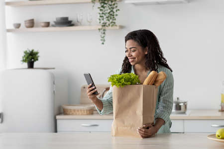 Delivering products to home. Smiling millennial african american female looks at smartphoneの写真素材