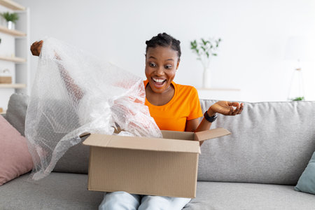 Express delivery service. Happy black lady receiving package, unpacking cardboard box, sitting on sofa at homeの写真素材