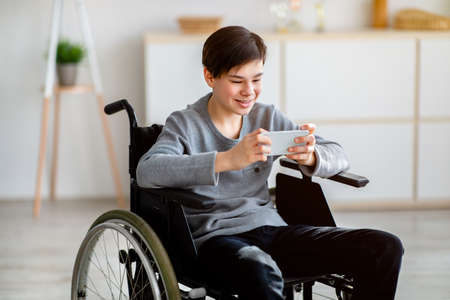 Portrait of cheerful handicapped teen boy in wheelchair using smartphone, chatting and texting in...