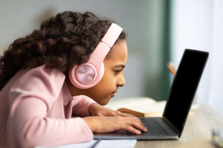 Youth and technology concept. Focused black schoolgirl using laptop computer, sitting at table too close to pc, side viewの写真素材