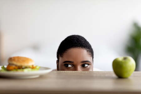 Overweight black woman choosing between burger and apple, peeking at food from under table, copy...