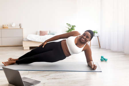 Online sports for weight loss. Overweight black woman doing exercises in front of laptop, standing...