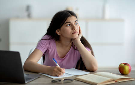 Thoughtful Indian teen girl studying online from home, taking notes during web lesson, feeling bored...