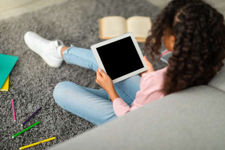 E-learning concept. African american schoolgirl using digital tablet with empty screen, sitting on floor with booksの写真素材
