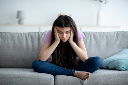 Sad Indian teenage girl sitting on sofa at home with head in hands, suffering from lack of communication and lonelinessの写真素材