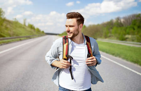 Handsome young man with backpack walking along highway, traveling by autostop, hitchhiking in...