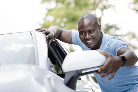 Cheerful black man standing by white car, looking at mirror