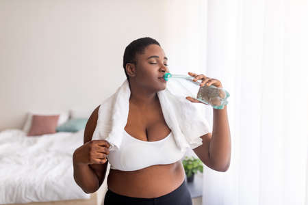 Healthy living, weight loss. Plus size black lady drinking fresh clear water from bottle after sports workout indoorsの写真素材