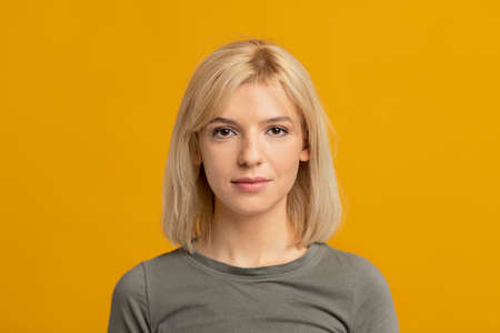 Portrait of confident serious young woman with blonde hair looking at camera and posing over yellow studio backgroundの写真素材