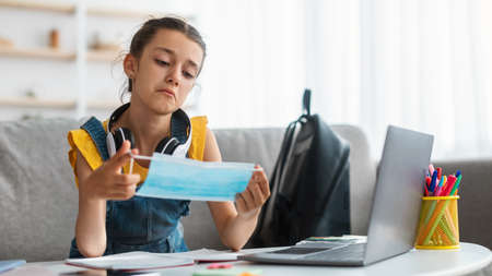 Sad girl holding medical mask studying at home with laptop