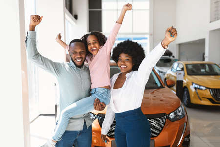 Excited black family of three lifting hands up, celebrating new car purchase in auto dealership