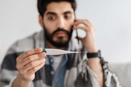 Shocked ill arab man calling doctor while looking at thermometer, sitting on sofa wrapped in...