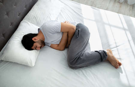 Stomachache. Young arab guy suffering abdominal pain touching aching stomach while lying in bed,...