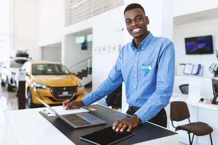 Portrait of happy car salesman standing at work desk, smiling at camera, using laptop and touch pad in auto dealershipの写真素材