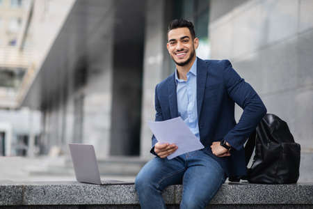 Handsome arab entrepreneur sitting by office building with laptopの写真素材