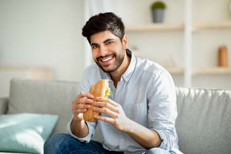Cheat meal day. Happy arab man eating tasty sandwich and smiling to camera, sitting on sofa in living room at homeの写真素材