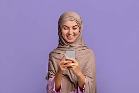 Cool application. Excited young muslim woman in hijab using smartphone, texting or browsing...