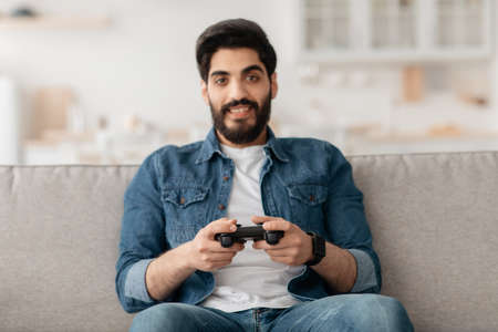 Relaxed middle-eastern guy playing video games with joystick at home, sitting on sofa, copy space