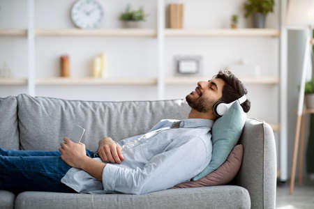 Happy arab guy in wireless headphones lying on couch with closed eyes and listening to music, copy...