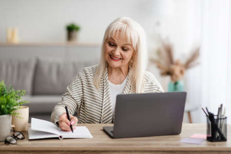 Smiling mature lady writing in notebook using laptop at homeの写真素材