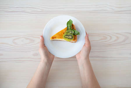 Delicious dessert. Woman holding plate with slice of cheesecake decorated with kiwi slices, wooden table, top viewの写真素材