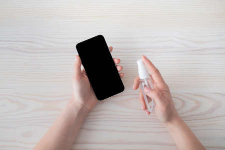 Gadget disinfection. Unrecognizable woman sanitizing smartphone with disinfectant mist spray, top...