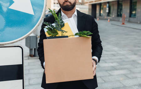 Unemployed businessman standing with box of stuff outdoors, near road sign, lost his job, cropの写真素材