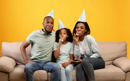 Happy african american parents and girl celebrating birthday and blowing at party horns over yellow backgroundの写真素材
