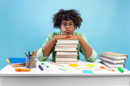 Lack of motivation. Bored african american teen guy sitting at table and leaning on stack of books, blue backgroundの写真素材
