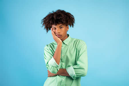Portrait of black teen guy feeling bored, leaning on his hand, having dull day on blue studio...