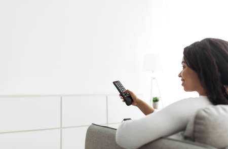 Young black lady holding and pointing remote control on white empty wall, mockup for your design or...