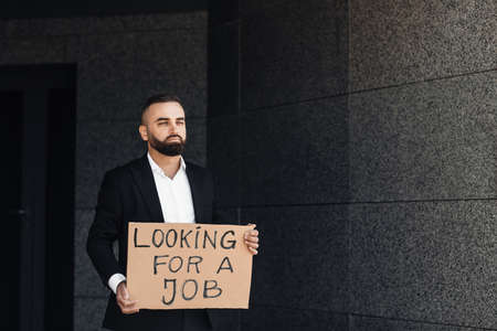 Unhappy despaired businessman lost work holding looking for job placard, standing near business...