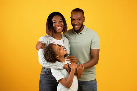 Happy family. Loving african american parents with daughter embracing and smiling at camera over yellow backgroundの写真素材