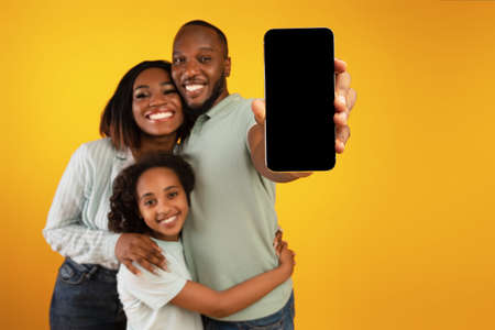 Loving african american family of three showing blank smartphone screen to camera, standing on...