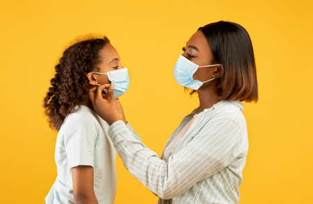 Loving african american woman putting medical mask on his cute daughter face, yellow studio backgroundの写真素材