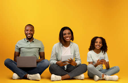 Family gadgets. Excited black parents and their pretty daughter using different electronic devices,...